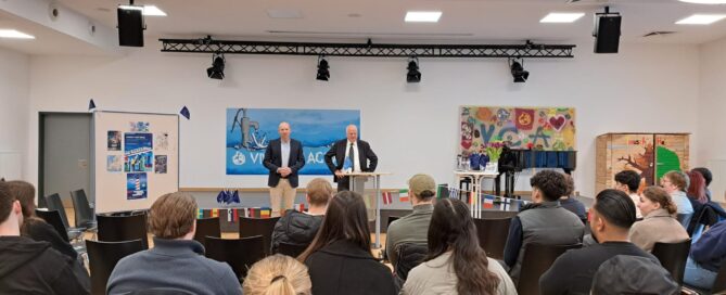 Zu sehen ist die Aula der Schule. Auf der Bühne stehen der Schulleiter und der Staatsrat.