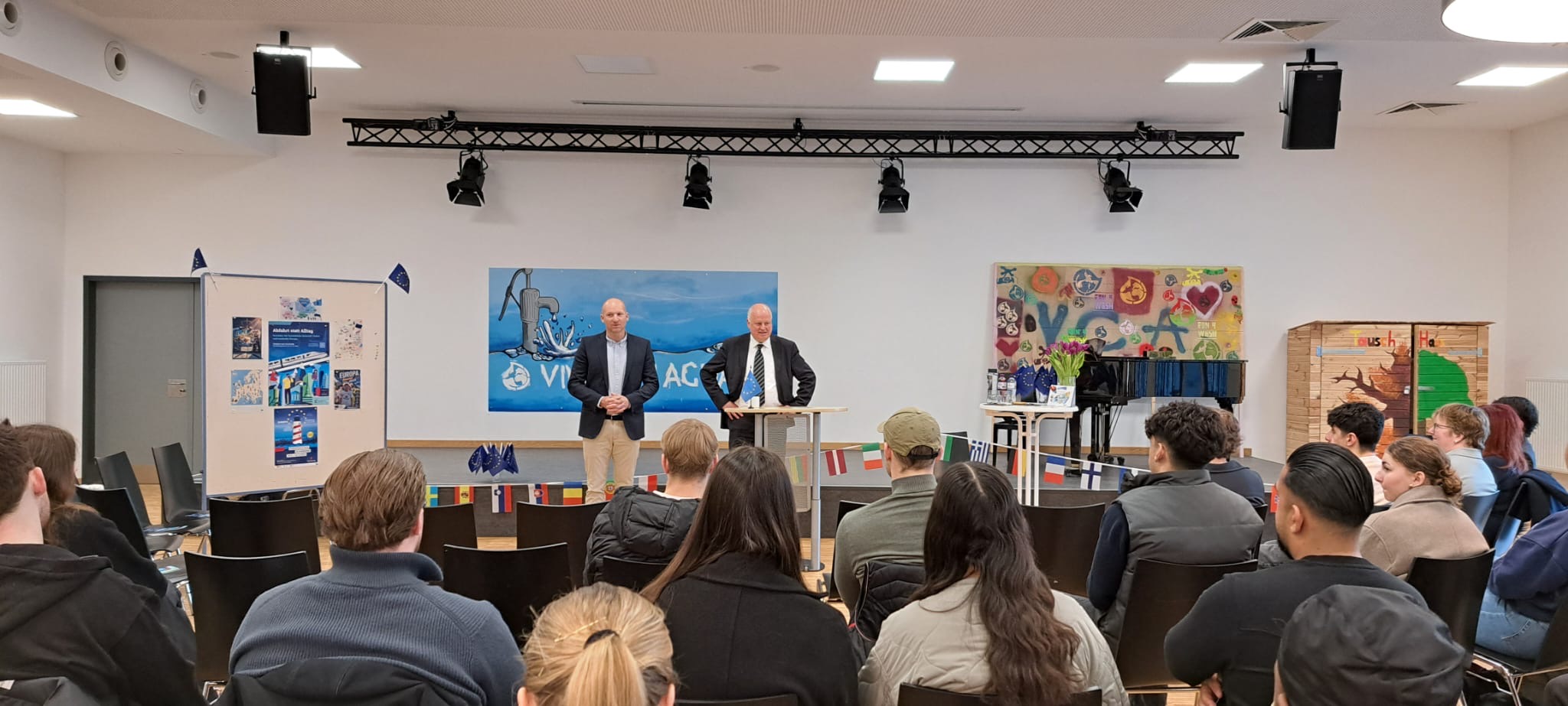 Zu sehen ist die Aula der Schule. Auf der Bühne stehen der Schulleiter und der Staatsrat.