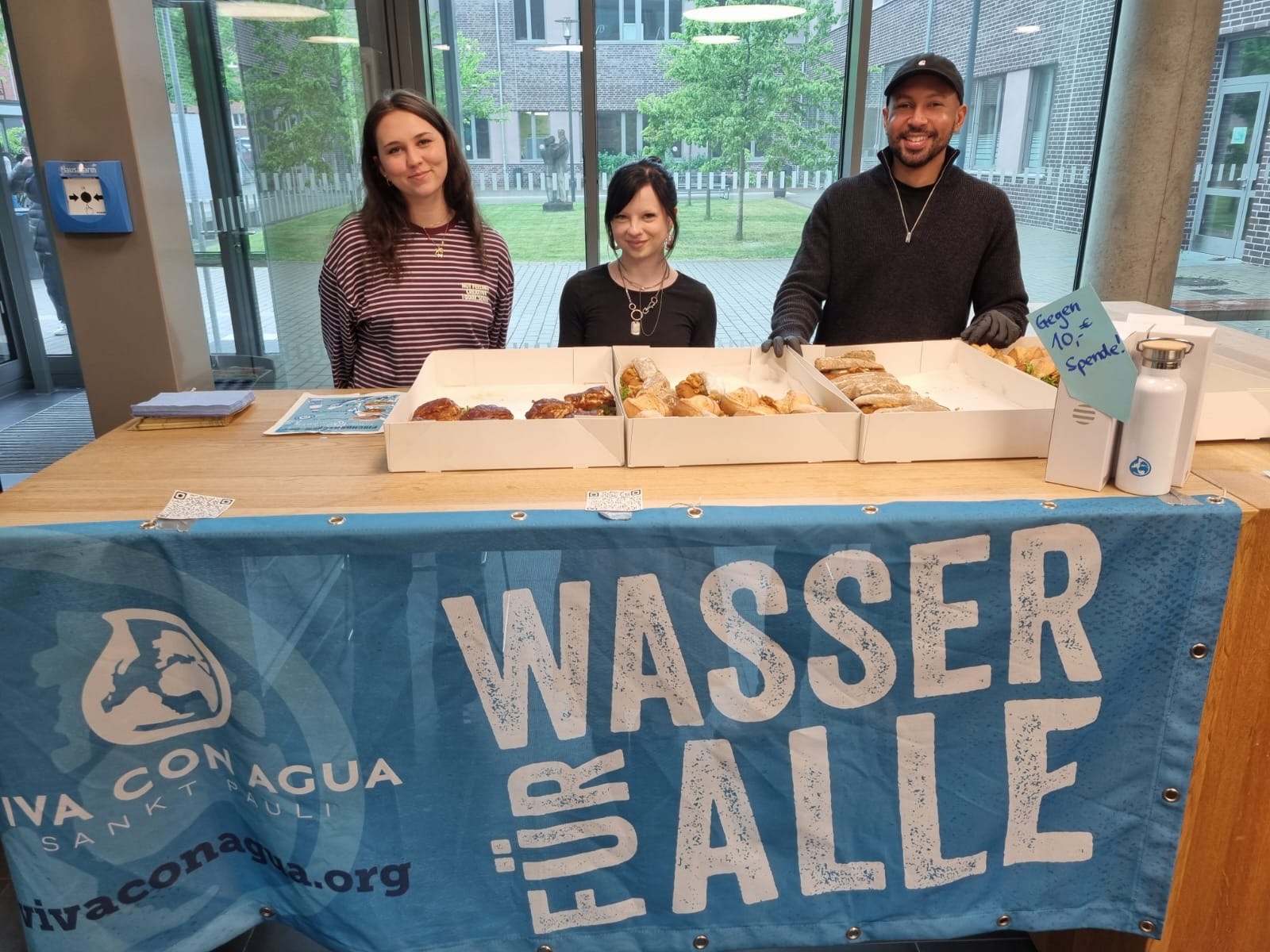 Zu sehen sind drei Schüler:innen, die Fischbrötchen im Foyer der Schule verkaufen. Sie stehen im Foyer.