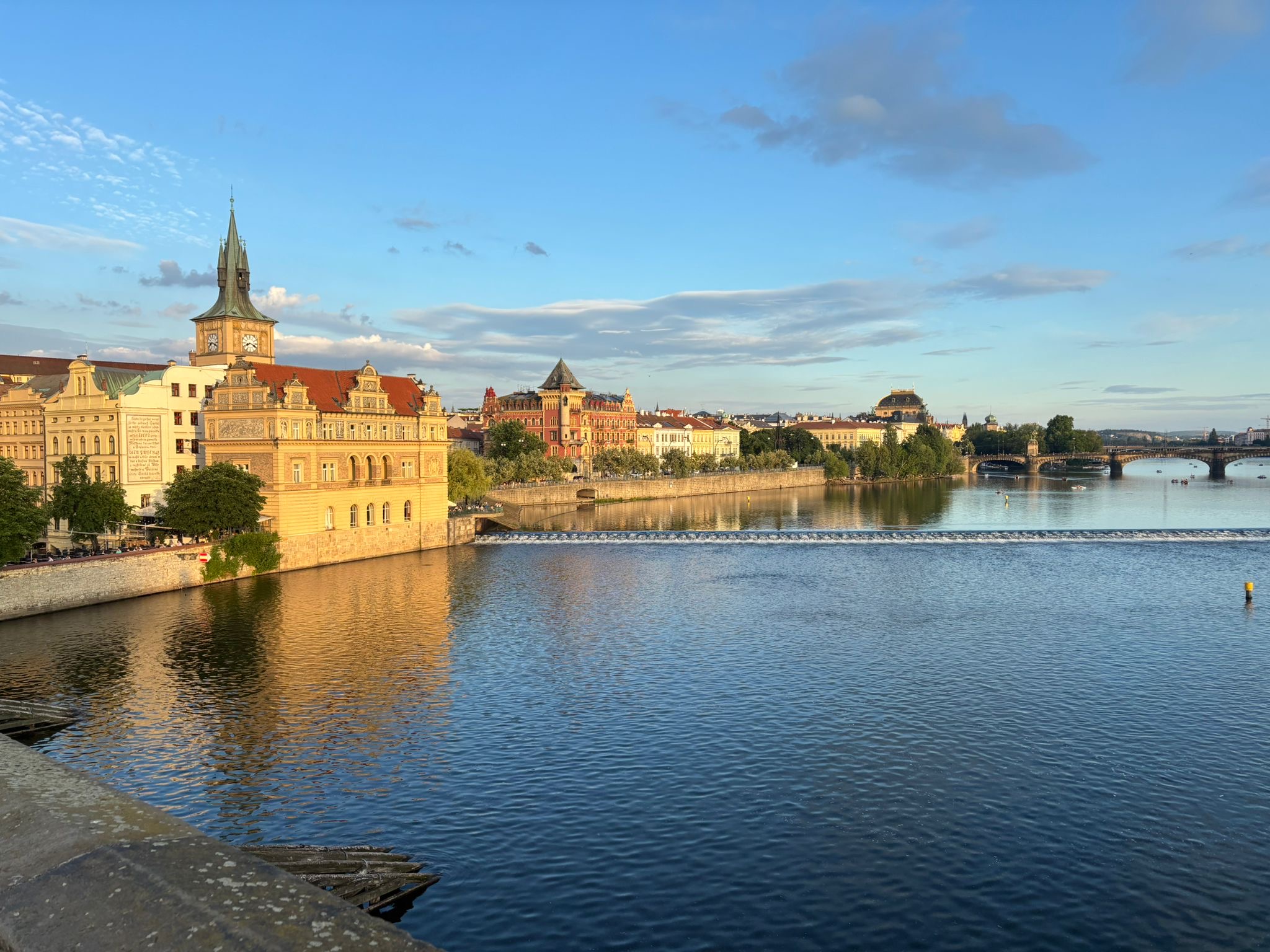 Auf dem Foto ist die Moldau und Gebäude an der Moldau und im Hintergrund die Prager Burg im Sonnenschein zu sehen