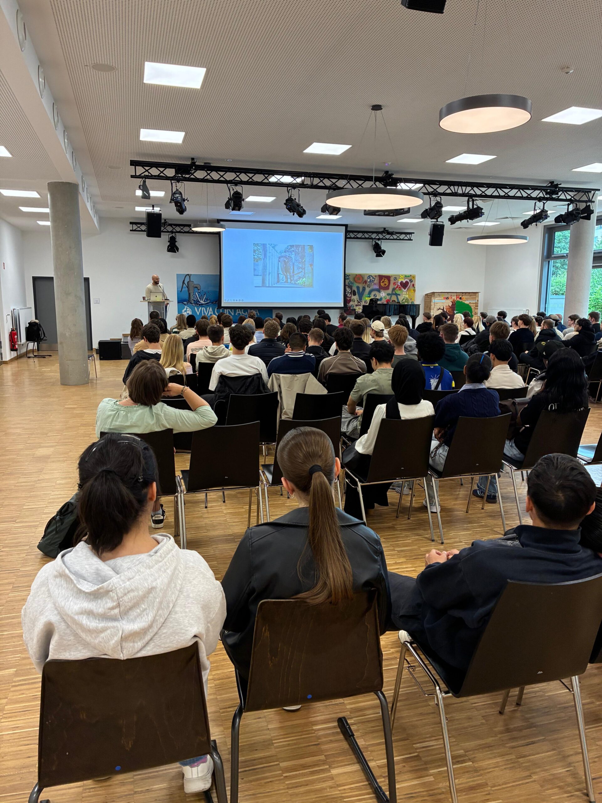 Blick von hinten in eine gut gefüllte Aula: Schülerinnen und Schüler sitzen auf Stühlen und hören einem Vortragenden zu, der vorne neben einer großen Leinwand steht. Auf der Leinwand ist eine Präsentation zu sehen.