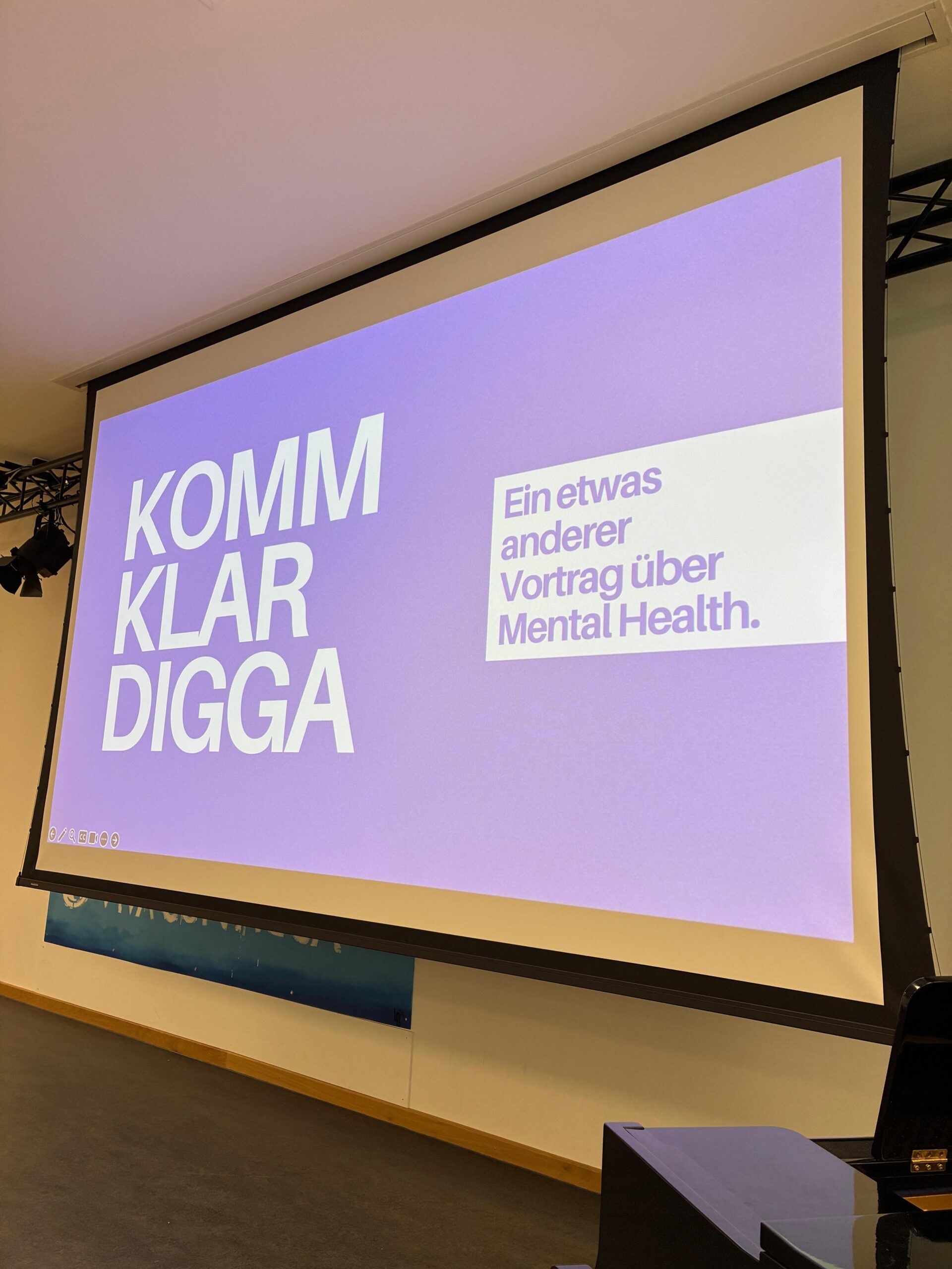 Große Leinwand in einer Aula mit einer lila Präsentationsfolie. Darauf steht links „Komm klar Digga“ und rechts „Ein etwas anderer Vortrag über Mental Health.“