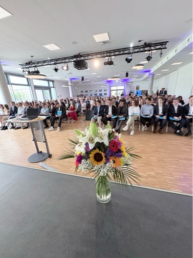 Blick in die Aula Blick von der Bühne in eine gut gefüllte Aula mit festlich gekleidetem Publikum; im Vordergrund steht ein großer Blumenstrauß.