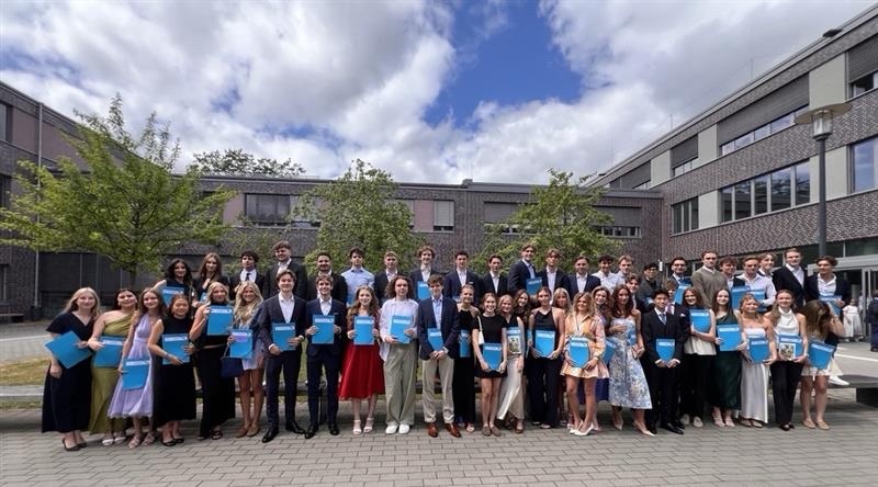 Große Gruppe von Absolventinnen und Absolventen steht mit blauen Urkundenmappen vor einem Schulgebäude und posiert für ein gemeinsames Gruppenfoto.
