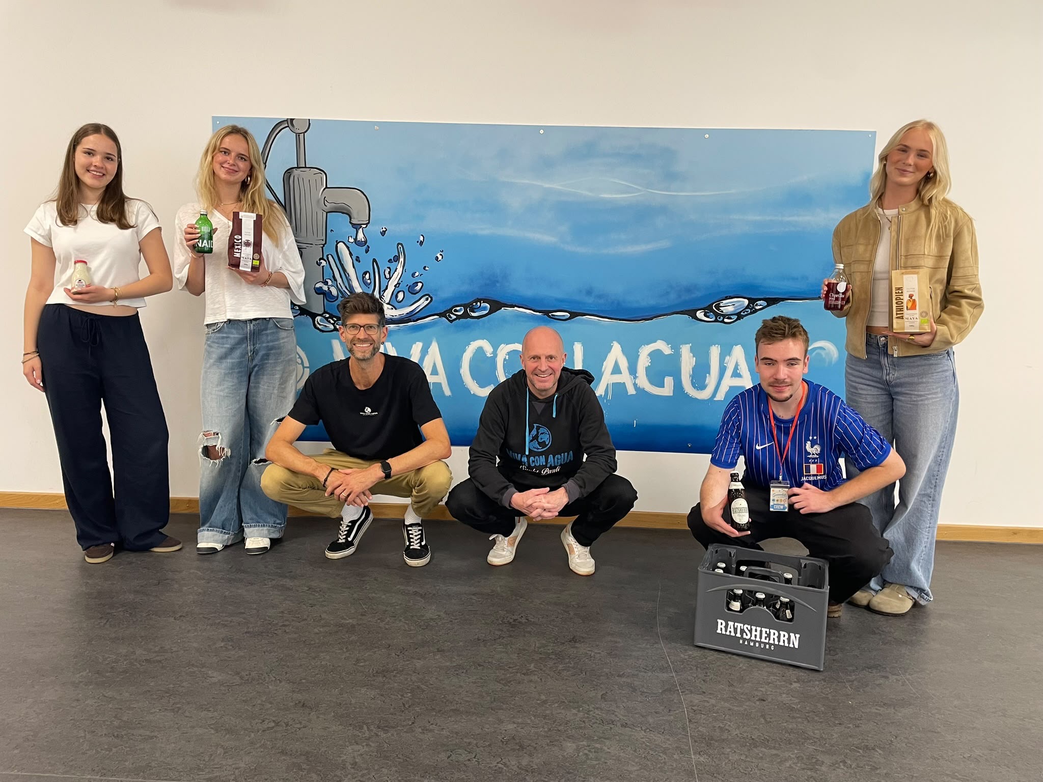 Zu sehen sind 6 Personen auf der Bühne in der Aula. Sie stehen vor einem Viva con Agua Bild und grinsen in die Kamera